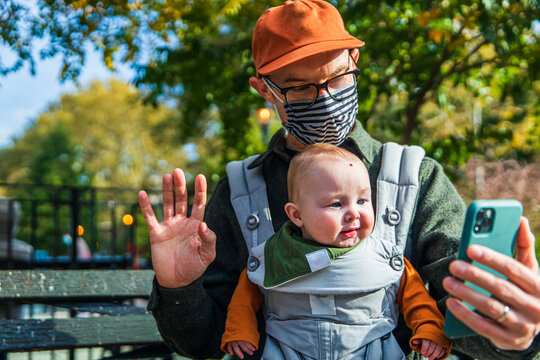 Father with cute baby daughter video calling through mobile phone while sitting on bench during COVID-19
