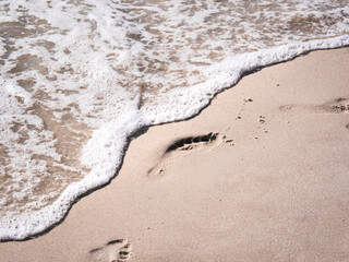 footprints in the sand along a beach with diagonal white foam of a wave for the concept of travel or remember loved ones