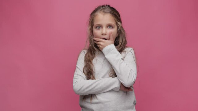 Portrait of surprised little girl at pink background. Cute curly-haired girl opening eyes and looking at camera. Surprised and shocked face expression