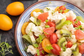Vegetarian salad of cauliflower cabbage, kiwi, tomatoes, microgreen sprouts on black concrete background. Side view, selective focus.