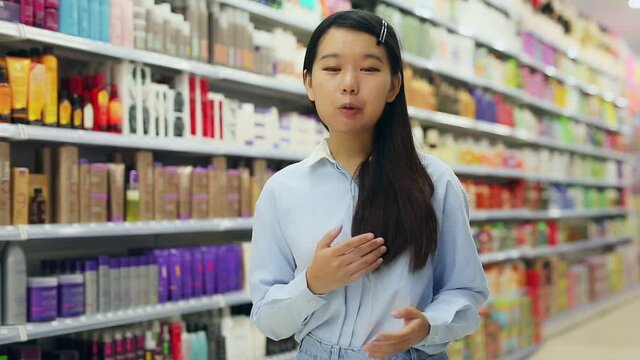 Portrait of friendly Asian female shop assistant near shelves with hair dye in cosmetics shop