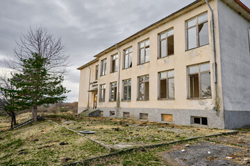 07.01.2021. Bulgaria, Kardzali. Old brownfield and abandoned soviet type school with overcast and very cloudy sky. School inside the green grass with broken windows.