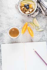 Empty opened white notebook with a pencil over it, a granola bowl at the top and a foamy coffee cup with some orange and apple slices in the middle.