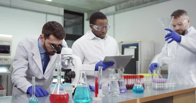 Portrait of male mixed-races microbiology scientists working in modern medical laboratory conducting genetic experiment using professional microscope equipment examining vaccine sample writing results