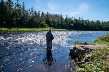 A male salmon angler stands in a river up to his waist with a long fishing rod wearing waders, fly hat and screen. The background is trees and a riverbank. The blue water has a small ripple on top