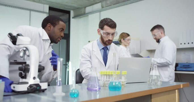 Portrait of busy African American male lab specialist in hospital laboratory looking in microscope doing medical research speaking with Caucasian colleague who is typing on laptop, developing cure