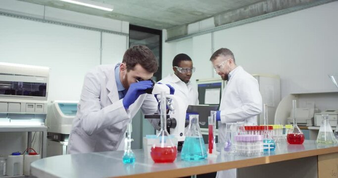 Caucasian male scientist specialist in laboratory working on microscope developing virus disease cure, medical research. African American doctor discussing analysis with co-worker behind. Portrait