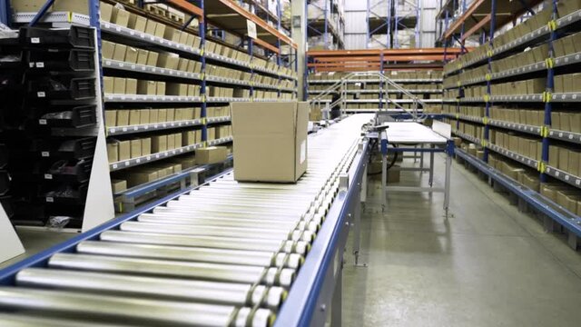 Traveling shot follows a cardboard box moving on long straight roller conveyor in large warehouse.