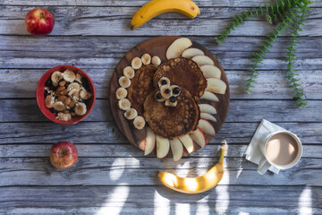 desayuno por la mañana un plato de madera con hot cakes con rebanadas de manzana y bananas alrededor con un tazon de frutas con una taza de cafe 