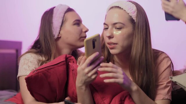 Four happy pretty young women in pajamas, Eye Collagen Patches and headbands on head sits on bed and uses phones at bachelorette party