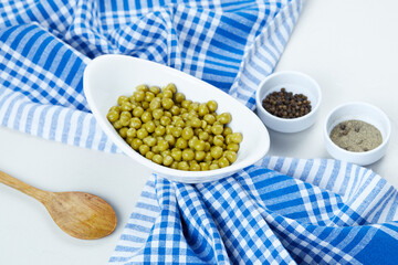 Boiled green peas in a white bowl on a white background with spices, a spoon, and a tablecloth