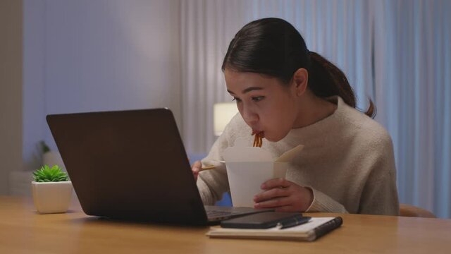 Young attractive beautiful asian female hungry eat instant noodles chinese food box take away full mouth look at computer notebook at home in busy work from home late night multitask unhealthy meal.