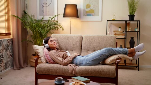 Calm Young Smiling Caucasian Woman Wearing Headphones Lying And Relaxing With Eyes Closed On Sofa In Home, Listening To Favorite Chill Music Enjoy Lazy Free Day Alone Concept Of Leisure And Spend Time
