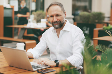 Adult male businessman sitting on the street in a cafe, drinking coffee and working on a laptop