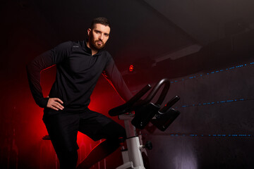 Fototapeta na wymiar male sitting on bike in gym posing at camera, workout ride a bike in gym, with the intention of healthcare. red neon smoky space in the background