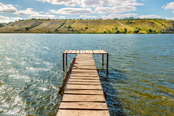 Obraz premium Wooden pier for fishing and swimming on bank of sunny river with hill slope on other side