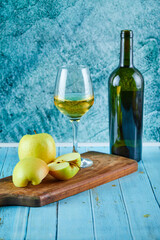 A glass of white wine and bottle with apple slices on blue background