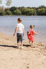 a little girl in a red dress and a boy are walking busik along the lake shore on a summer sunny day