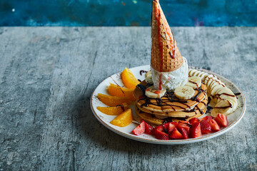 Pancakes with ice-cream cone, tangerine, strawberry, banana and chocolate syrup in the white plate...