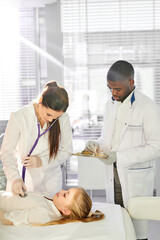 Fototapeta premium caucasian female doctor check the heartbeat of patient child with stethoscope while his african male colleague help her, in modern clinic