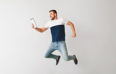 Young man with tablet computer jumping on light background