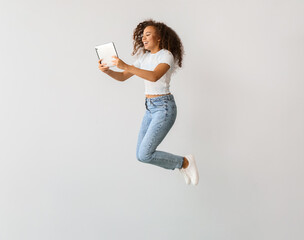 Young woman with tablet computer jumping on light background