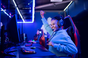 Streamer african young woman rejoices in victory professional gamer playing online games computer with headphones, neon color - obrazy, fototapety, plakaty