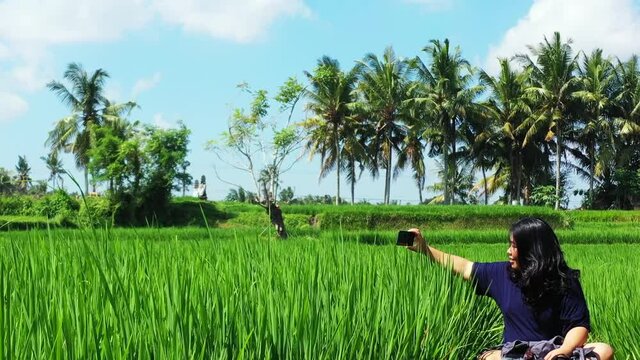 An Asian tourist smiling as she has fun relaxing in the middle of the bountiful fields of grass crops while recording the beautiful sights of the countryside, zooming in.