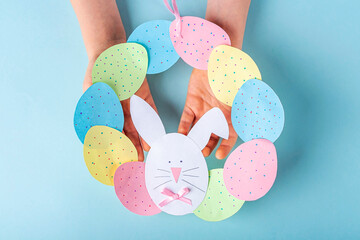 DIY and kids creativity. Step by step instruction: how to make paper easter wreath. Step11 Childrens hands holding finished cute wreath of paper eggs and bunny. Handmade Easter craft