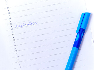 Close up view photo of word vaccination written in daily planner and blue pen on white background. To do list for day on white table. Vaccination flu, measles, coronavirus, covid-19 concept for people