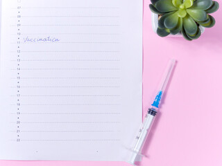 Vaccination concept. Close up view photo of word vaccination written in daily planner, syringe and succulent on pink background. To do list for day on pink table with office plant.