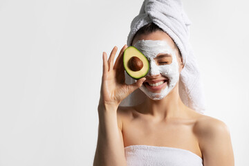 Overjoyed young pretty female with mask on face in white towel cover eyes by avocado.