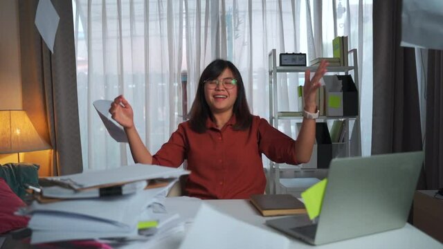 A young asian adult business woman sitting indoor room home office throwing papers documents notes fly in the air after taking a long time to complete the projects with happiness satisfied achievement