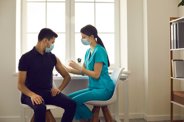 Side view of female nurse giving flu shot to male patient during vaccination campaign at the hospital. Doctor in face mask injecting young man with influenza, AIDS or Covid-19 vaccine at clinic office