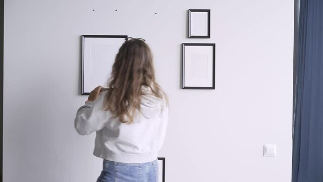 Young woman with long loose flowing curly hair hangs different picture frames of various size on wall decorating home under bright light backside view
