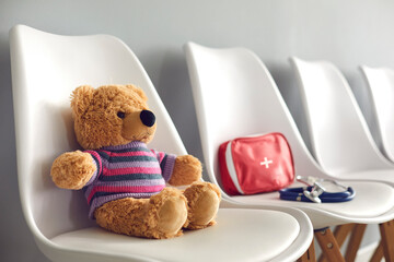First-aid kit, stethoscope and cute teddy bear on white chairs in waiting room of children's...