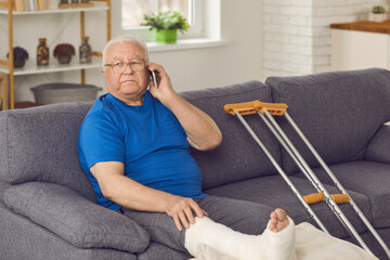 Serious senior man with broken leg in cast sitting on couch at home and making phone call on...