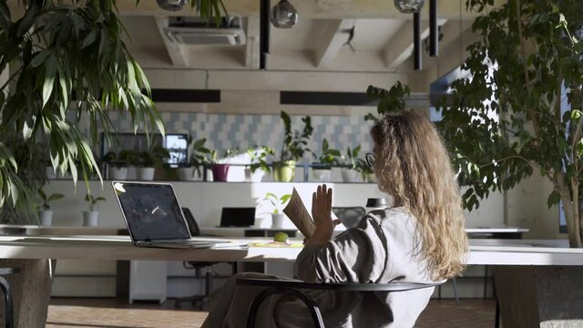 Professional woman lecturer with long loose curly hair holds notes and conducts online lesson sitting at table with laptop at home against pot plants