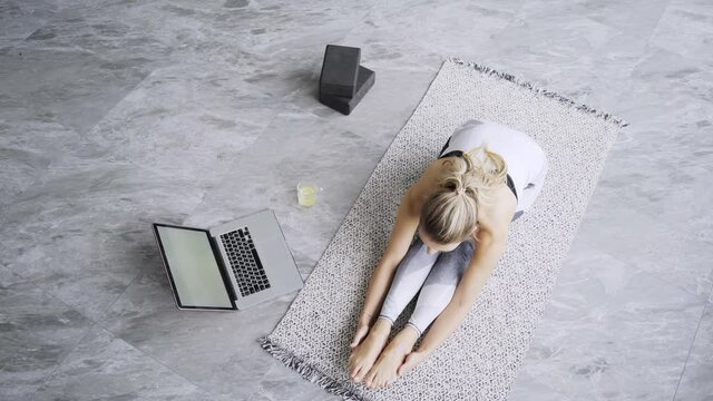 Professional yoga trainer in sportswear stretches back sitting on special carpet near grey laptop at home under bright morning light upper view