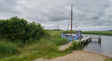 Nymindegab, Denmark - June 20, 2020: vintage blue fishing boat on a  jetty behind fresh green shore - the sky is cloudy