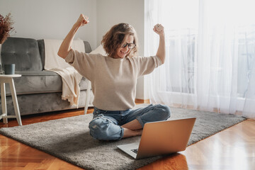Pretty curly happy young woman sitting on floor at home in front of laptop with victory gesture...