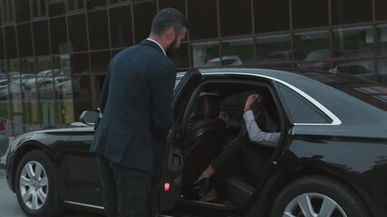 Medium shot of male driver in business suit opening door for successful businesswoman sitting in backseat of expensive black car
