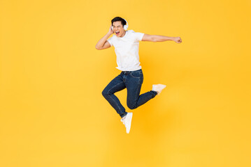 Happy handsome young Asian man wearing headphones listening to music and jumping in colorful yellow background