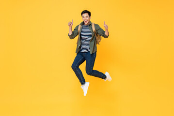 Fototapeta na wymiar Happy smiling young Asian tourist man with backpack jumping and giving thumbs up isolated on yellow background