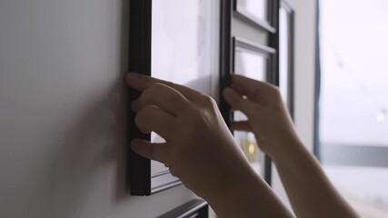 Person hands hang brown wooden frame with painting on wall among different pictures of various size against blurred window in new flat close view