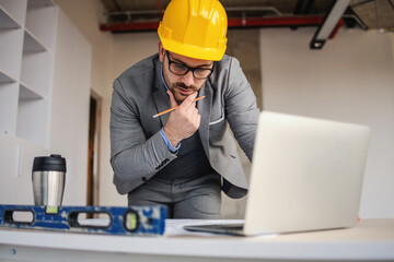 Focused architect looking at blueprints and thinking while leaning on desk at construction site.