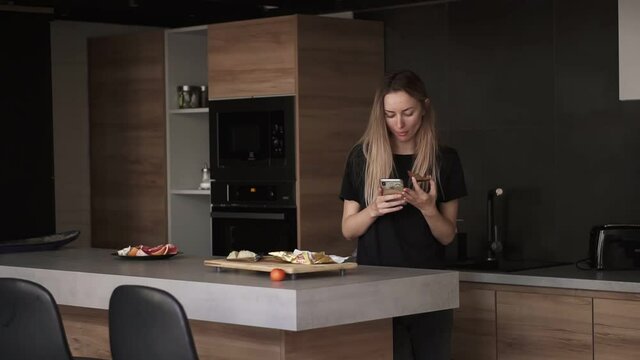 Concentrated woman eating bread with butter for breakfast, checking her smartphone in kitchen