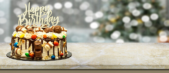 Birthday layer cake decorated with chocolate pieces with happy birthday text on marble table in front of abstract blurred Christmas bokeh background