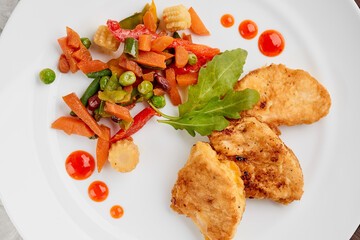 stewed vegetables with herbs and chicken steaks in batter on a white plate with drops of ketchup on a gray concrete background top view, food menu with a place to copy the text