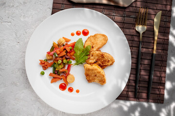 stewed vegetables with herbs and chicken steaks in batter on a white plate with drops of ketchup on a gray concrete background top view, food menu. fork and knife on a brown bamboo mat